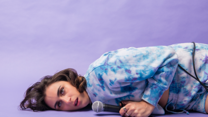 Image Description: A lilac background is behind Ashley, who lies on the floor on her side holding a wired microphone to the ground. Wearing a blue and white tie dyed outfit, her face is smooshed against the floor with an exhausted look on her face